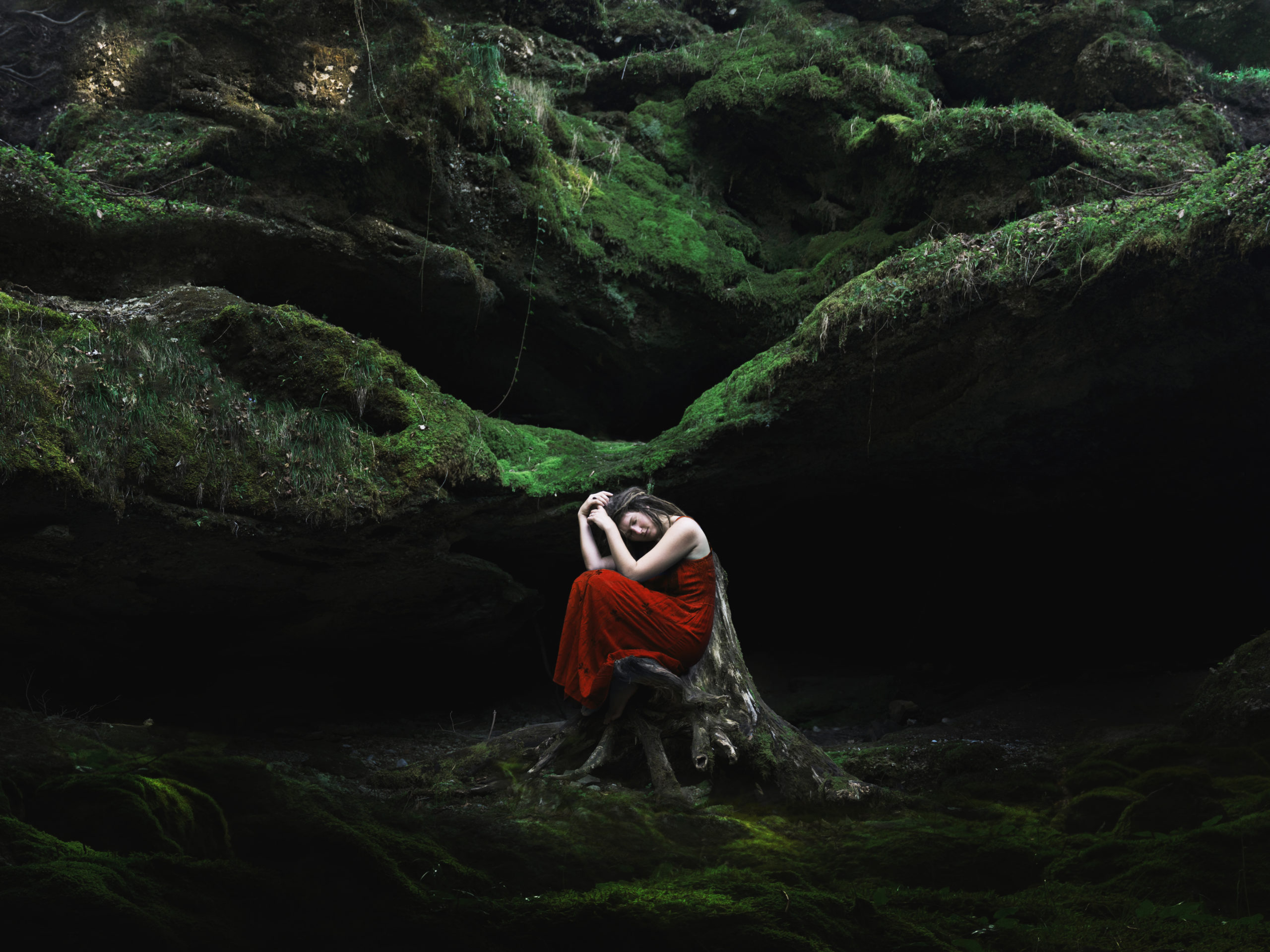 Self portrait showing a girl in a red dress in a contemplative pose in a green, mossy forest.