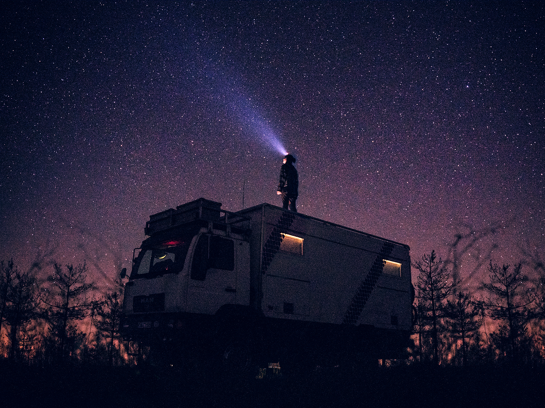 A person standing on an expedition vehicle shining a torch into the sky full of stars.