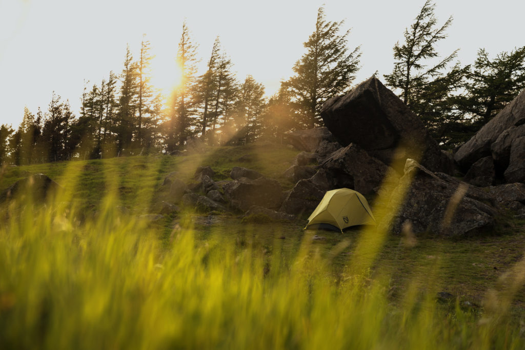 A green tent in the grass, sheltered by some large rocks, in the last sun of the day.