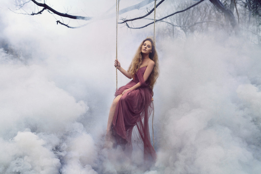 A self portrait of a woman in a long dress sitting on a swing surrounded by clouds.