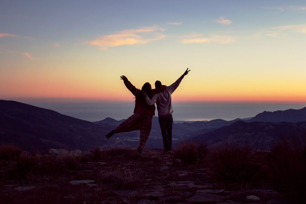 Two friends traveling together, depicted joyfully in a mountain landscape at sunset.