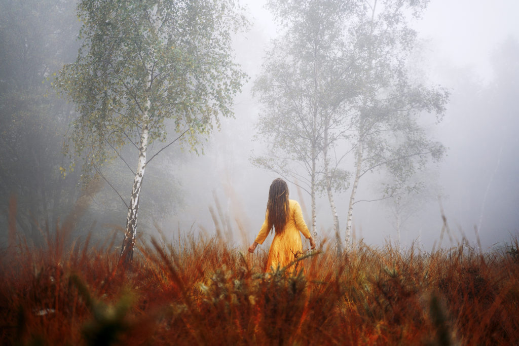 A creative self portrait of a woman standing in a field, surrounded by fog and trees.