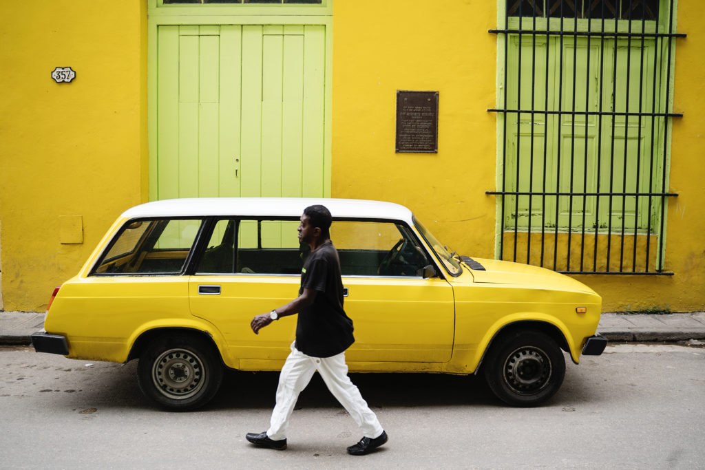A man walking in a colorful alley in Cuba. The walls as well as the car he walks past are bright yellow.