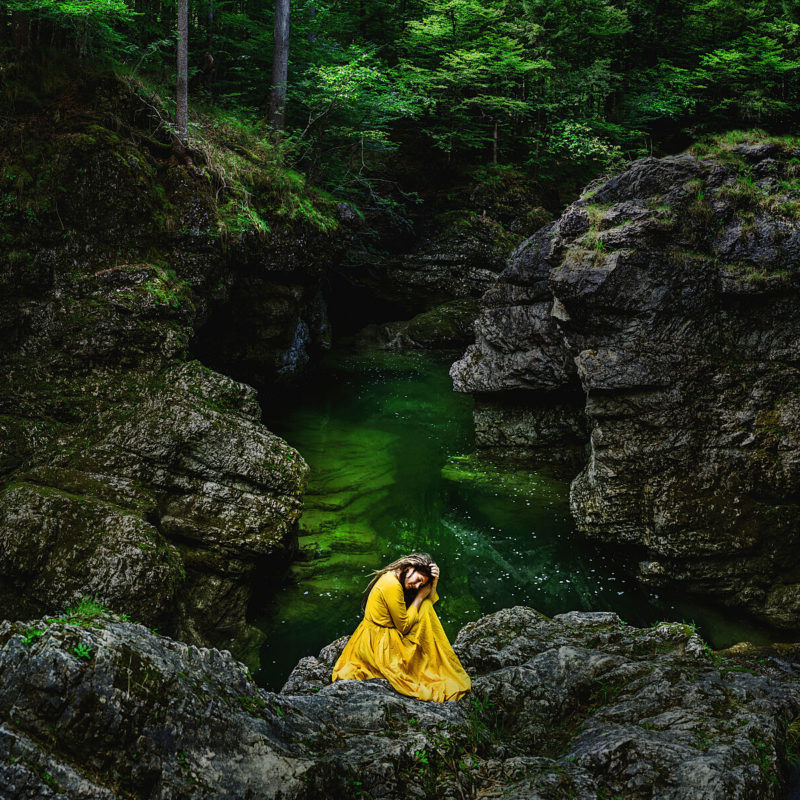 Tranquil, artistic self portrait photography in nature. The depicted woman wears a yellow dress which creates a beautiful contrast with the green environment of a gorge.
