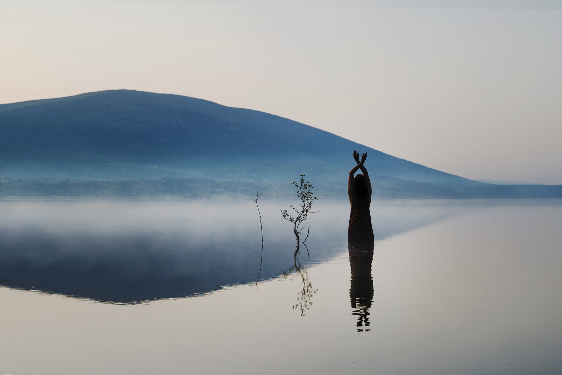 A Fine Art Photograph depicting a beautiful lake in the morning. The lake reflects the shapes of a woman and two branches. There is fog in the distance, creating a ethereal mood in this self portrait.