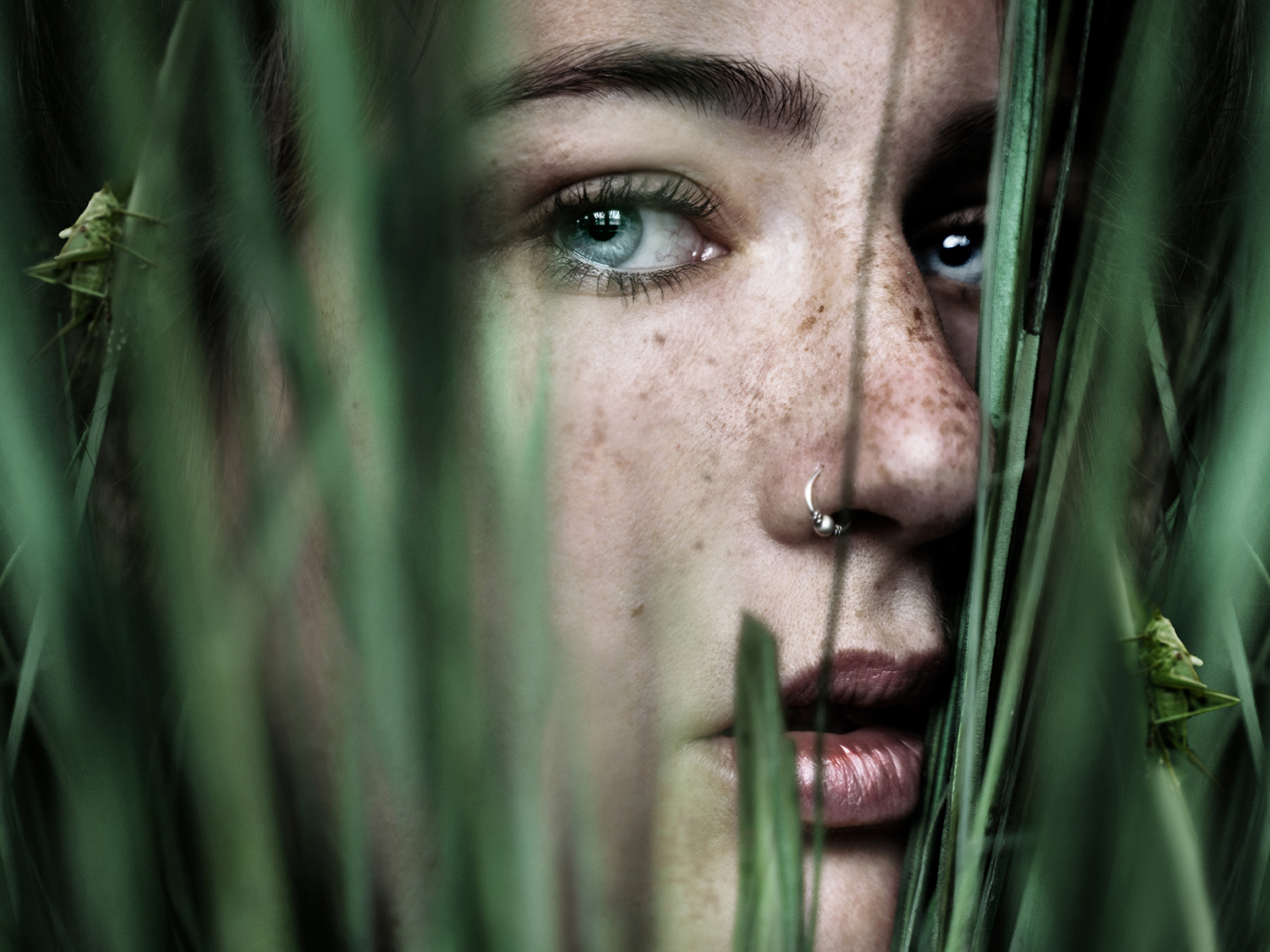 A beautiful close-up self-portrait of a woman with freckles. Her face is surrounded by grass and some hidden grasshoppers.