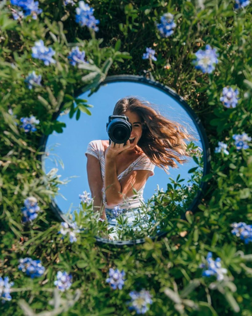 Mirror self-portrait photography is a fun way of capturing creative photos of yourself. An outdoor scene with a young woman capturing herself in a mirror lying on a flowery meadow.