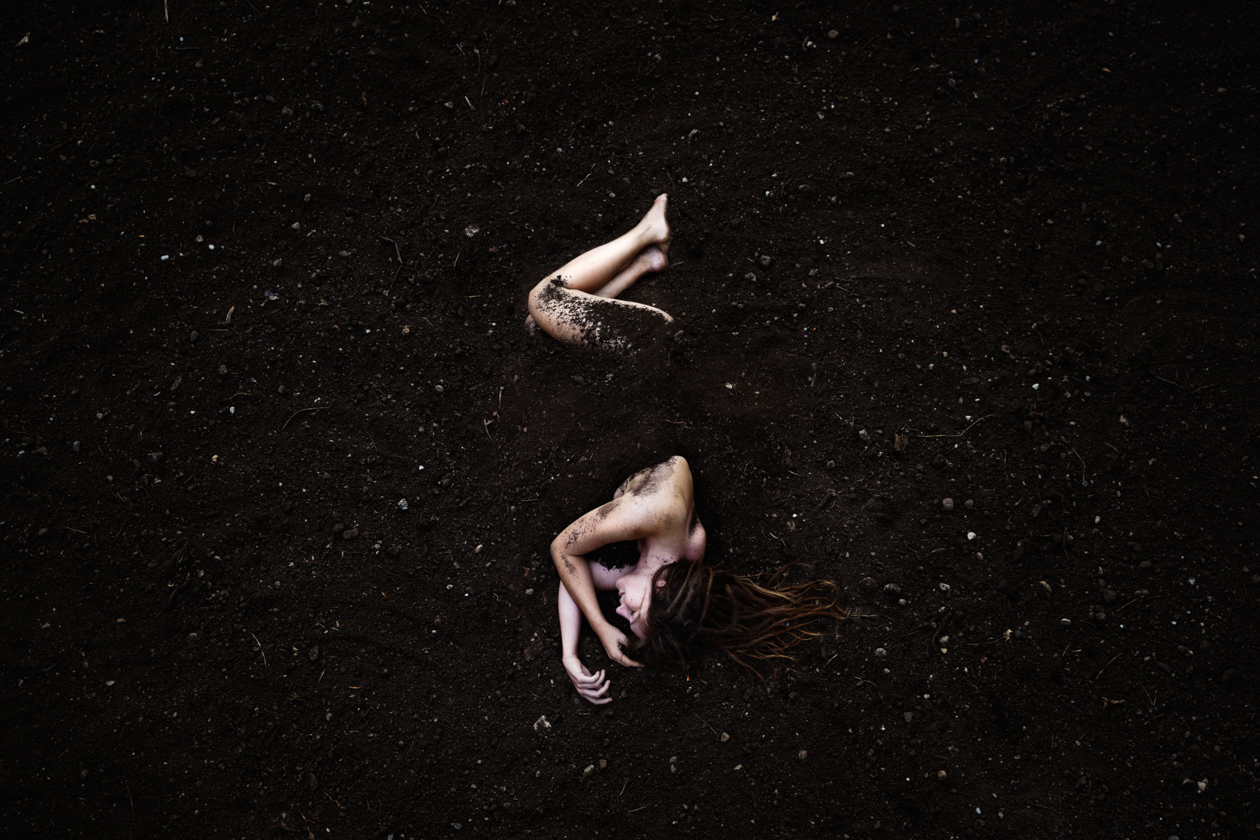 Mother earth self portrait photography, depicting a woman almost completely covered by soil, only her bare upper body and legs are visible among the dark surroundings.