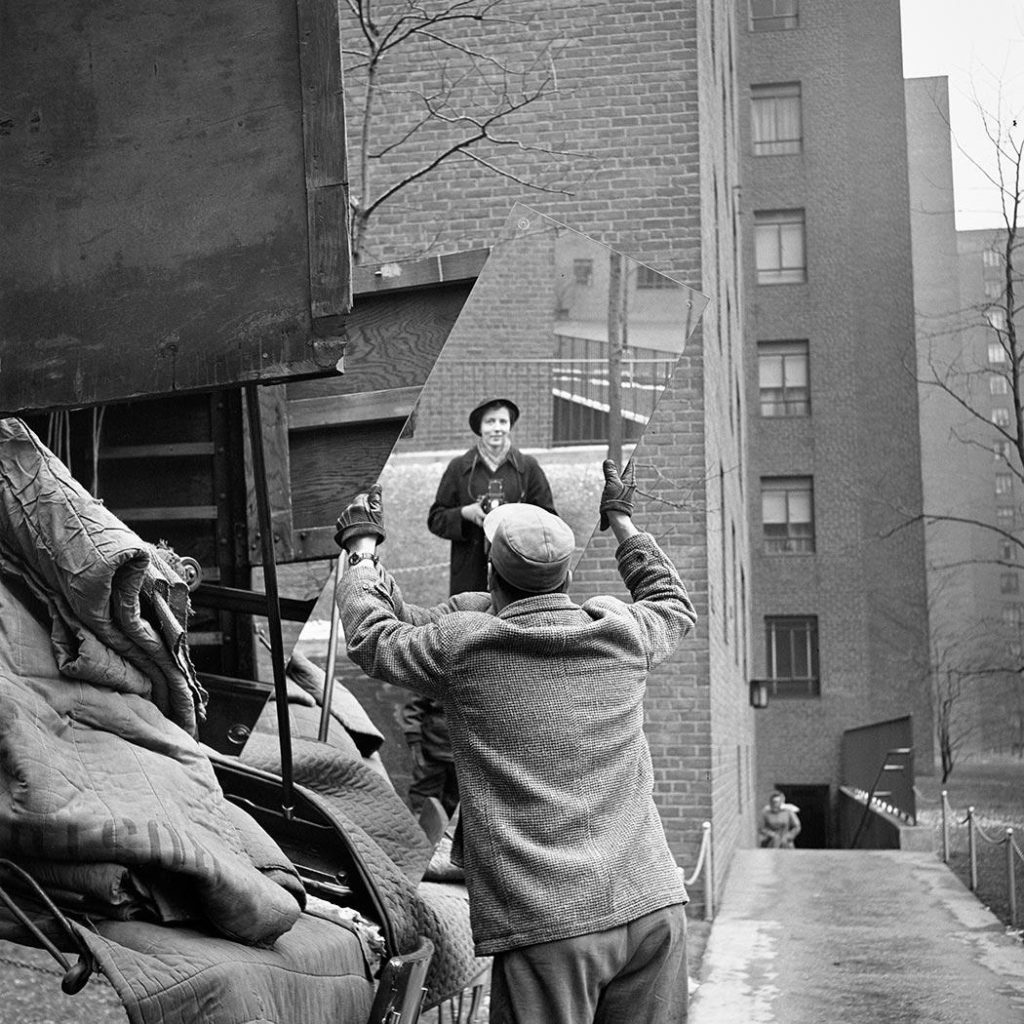 A famous black-and-white self-portrait by Vivian Maier. The photographer captures herself in a mirror, held up by a man in the streets. It seems like she just caught a moment of moving houses.